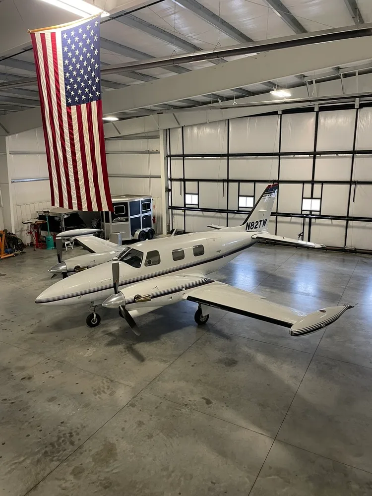 The white twin-engine aircraft N82TW with dark stripes parked inside a hangar, with a large American flag hanging overhead.