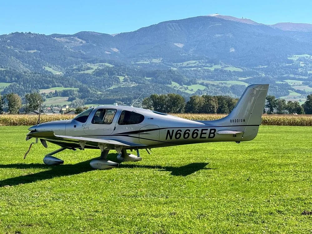 A silver Cirrus SR22 aircraft with black stripes is parked in a grassy field with mountains in the background.