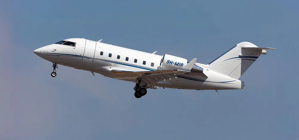 A white Bombardier Challenger 605 business jet with blue stripes, registration 9H-MIR, flying against a blue sky with landing gear extended.