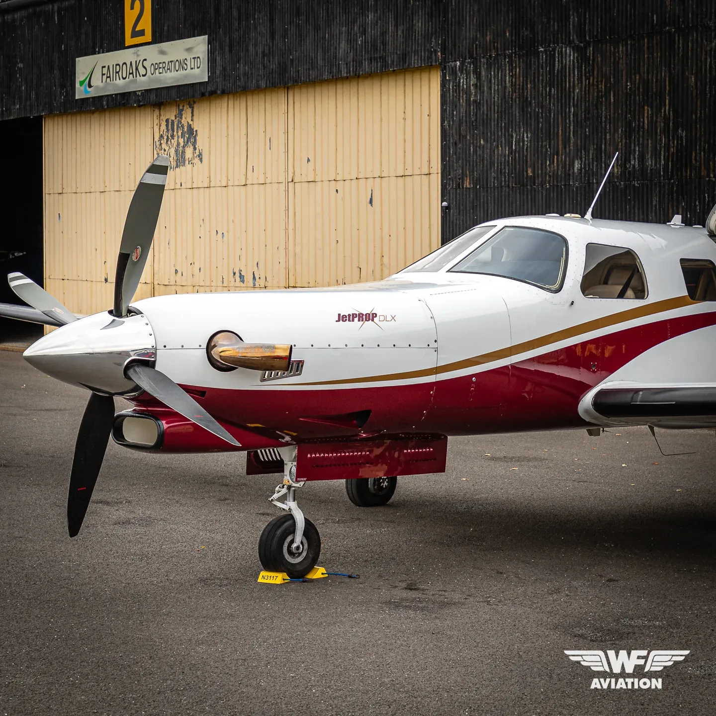 A white JetPROP DLX turboprop aircraft with red and gold stripes, registration N3117, parked on an tarmac in front of a hangar.