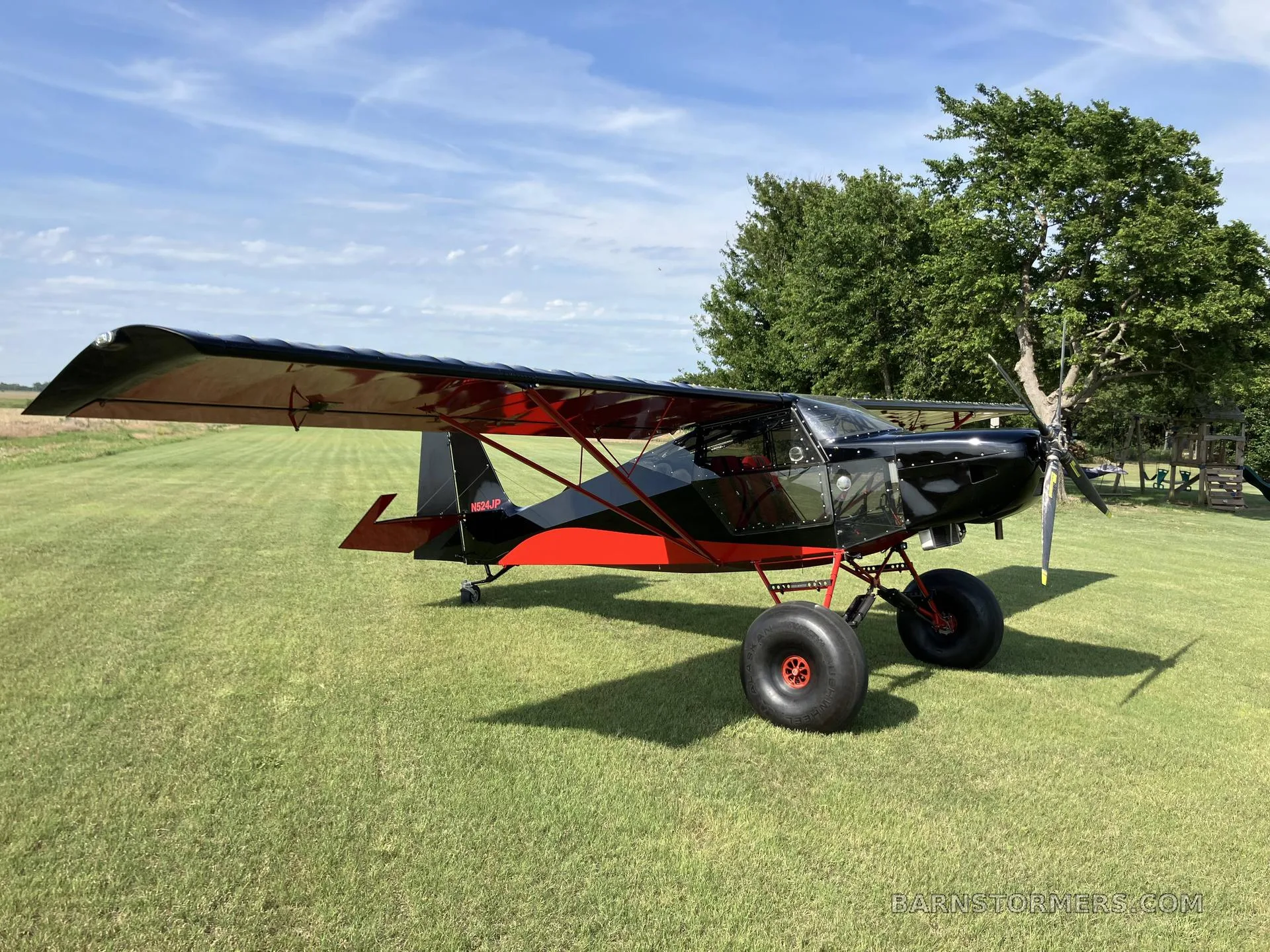 A black and red bush plane with large tires is parked on a grassy field, viewed from the front-right.