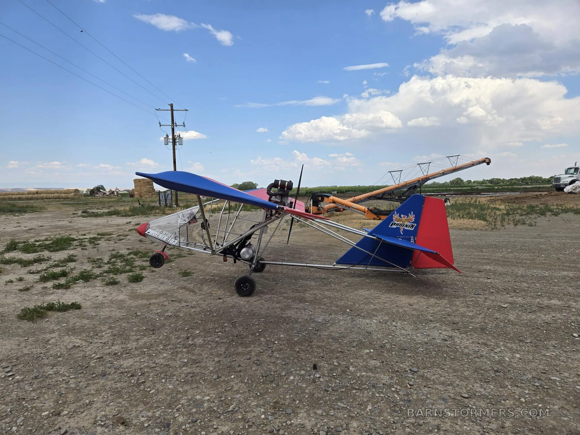 A red and blue ultralight aircraft with an exposed engine, parked on a dirt field under a partly cloudy sky.