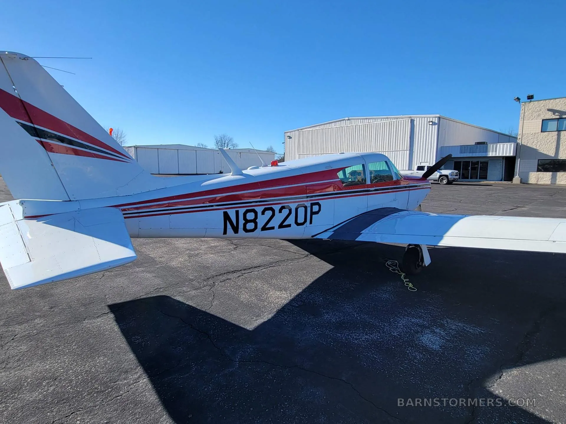 The tail and right side of a white and red Piper Comanche 260 (N8220P) are visible on an asphalt ramp with hangars in the background.