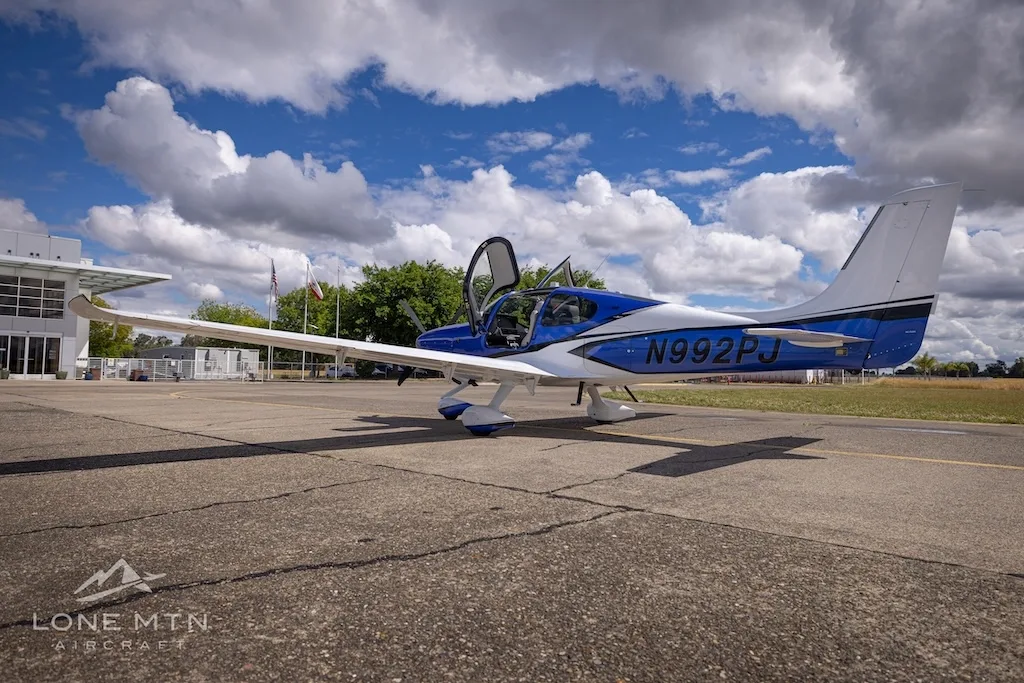 A blue and white Cirrus SR22 aircraft with its door open, parked on an airfield next to a building.