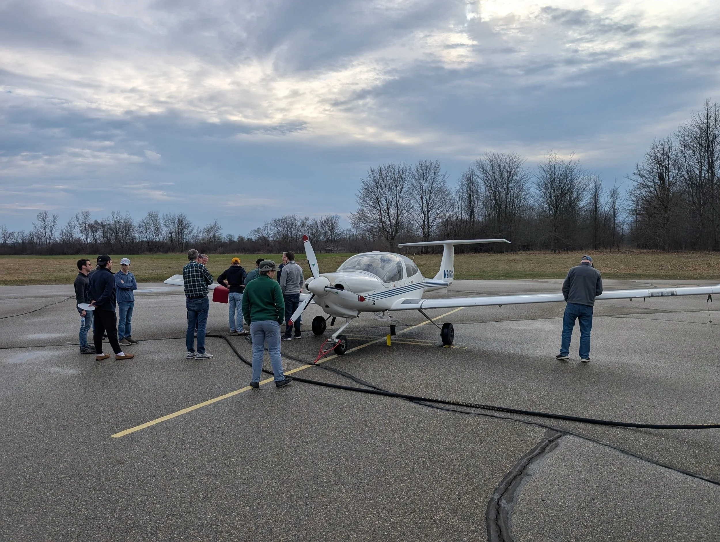 A white light aircraft with blue stripes on a tarmac, surrounded by people, under a cloudy sky.