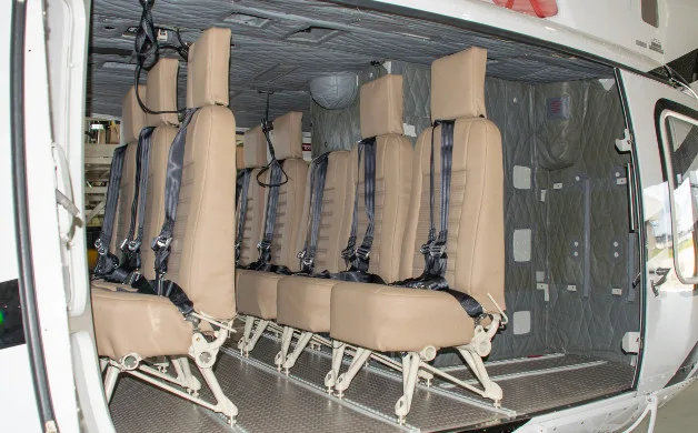 The interior of a helicopter cabin shows two rows of tan passenger seats with black harnesses.