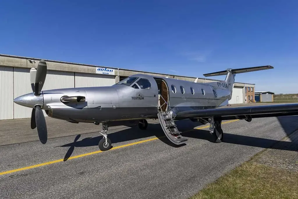 A silver Pilatus PC-12 aircraft with registration OY-GSA and its passenger door open, parked on an airfield.