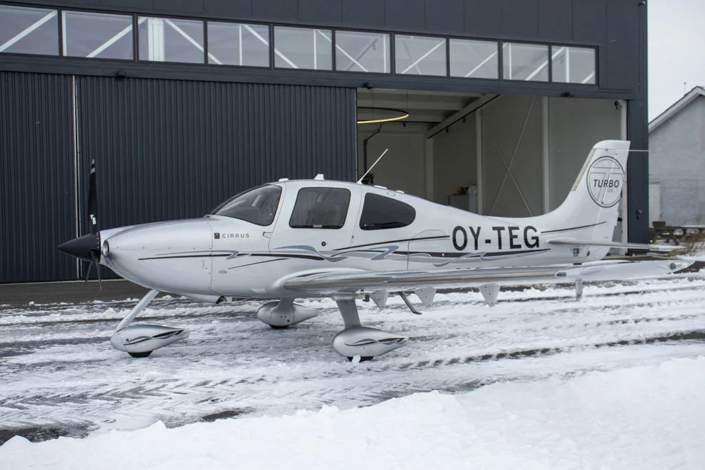 A silver Cirrus SR22 Turbo aircraft, registration OY-TEG, parked on a snowy ground outside a dark modern hangar.