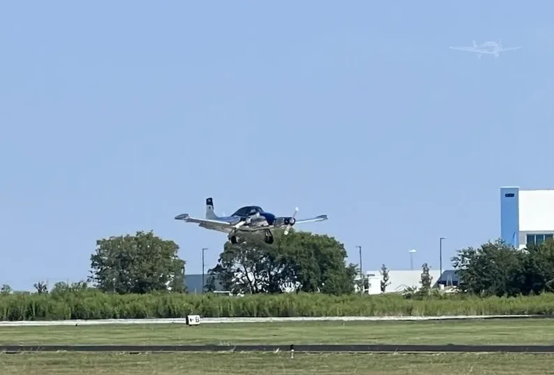 A silver and blue twin-engine aircraft, N708T, in flight near a runway with trees and buildings in the background.