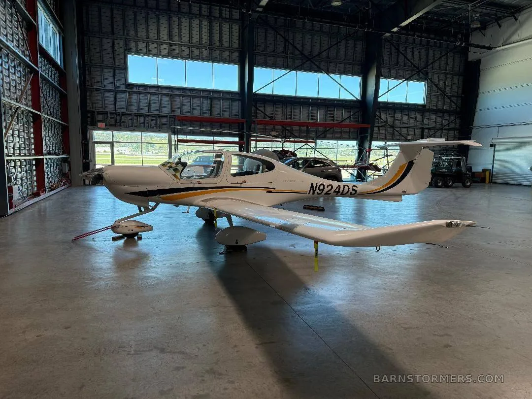 A white Diamond DA40 aircraft with yellow and black stripes, viewed from the side in a hangar.