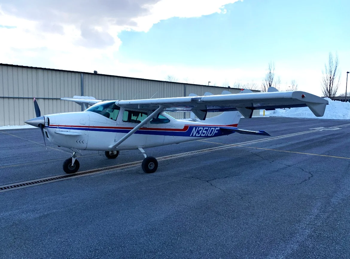 A white single-engine propeller aircraft with blue and red stripes (N351DF) parked on an airfield next to a hangar with snow in the background.