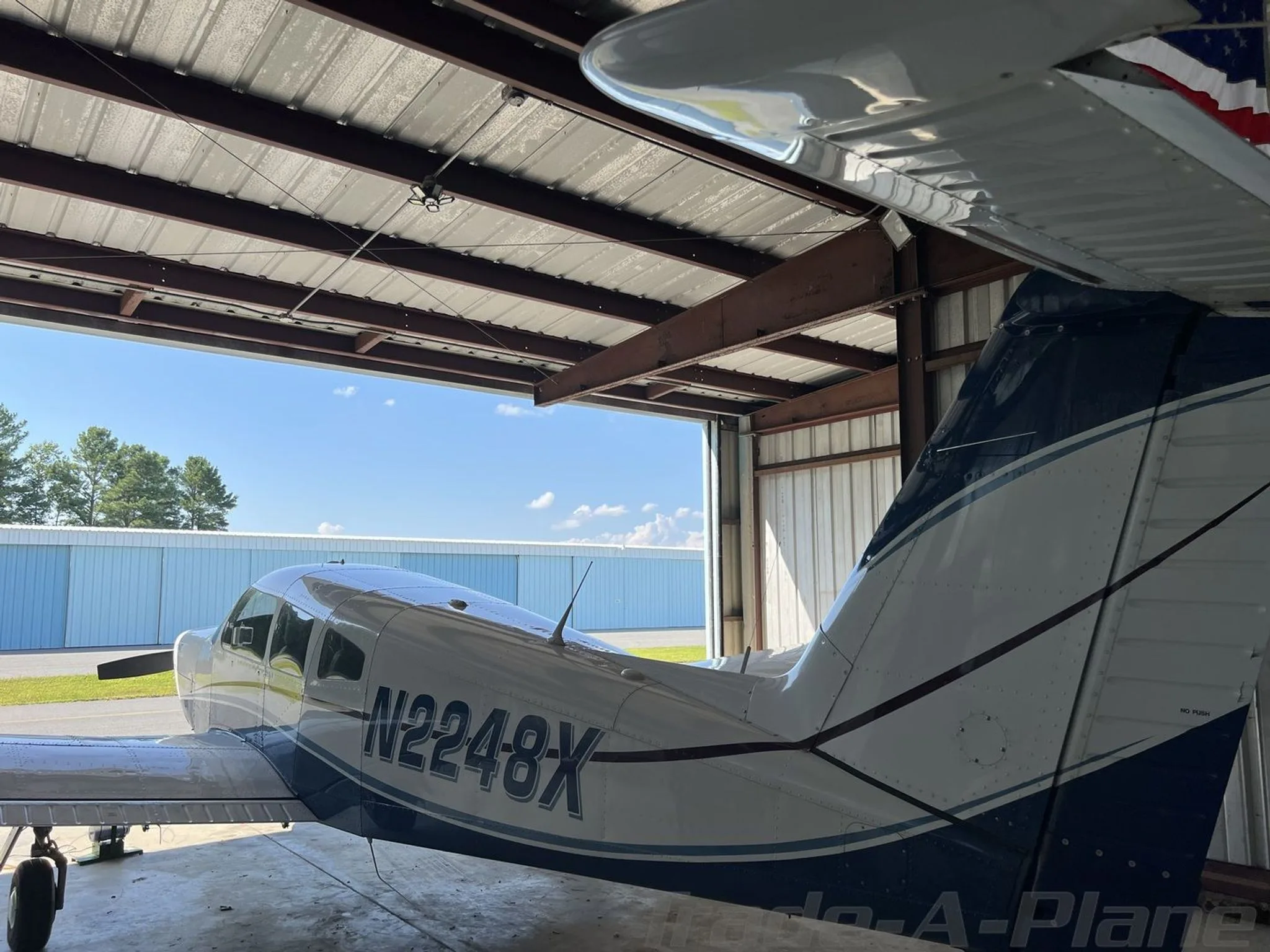 A view of the tail and fuselage of a white and blue Piper Archer II (N2248X) inside a hangar, looking out to a sunny airfield.