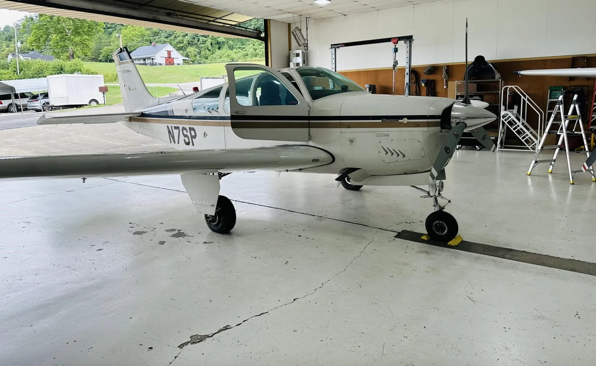 A white and gold Beechcraft Bonanza N7SP with an open door, parked in a hangar, viewed from the front right.