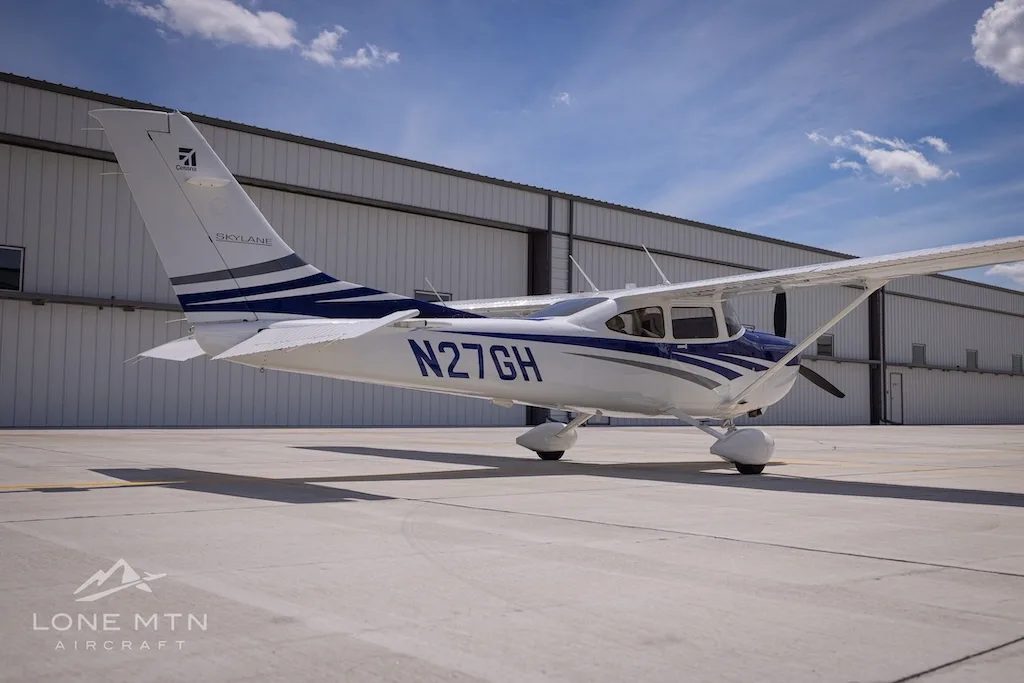 A rear-side view of a white and blue Cessna 182 Skylane (N27GH) parked on an airport tarmac.