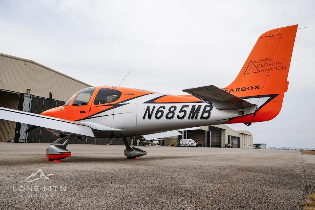 A side view of the orange and silver Cirrus SR20 aircraft N685MB parked near a hangar.