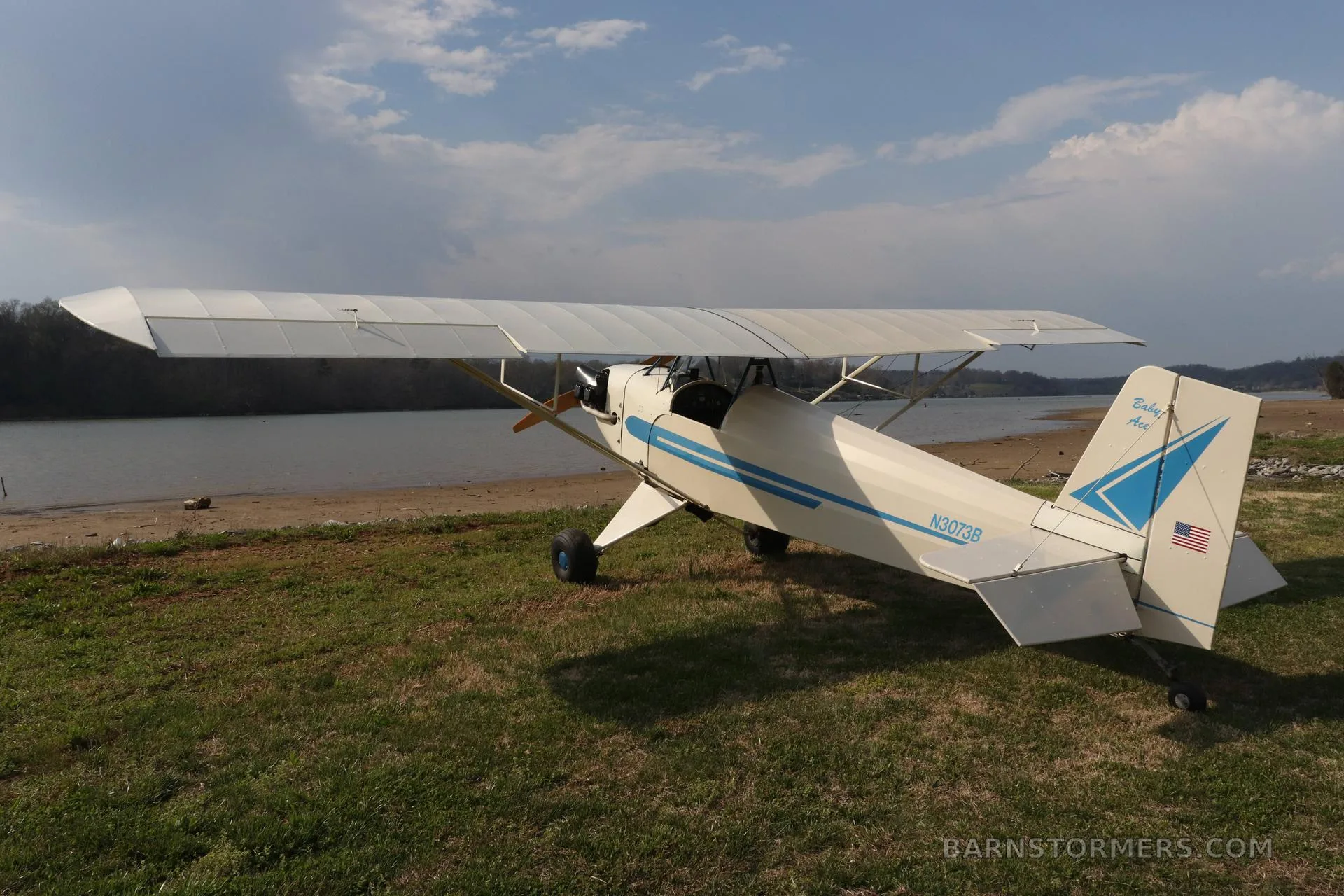 A white and blue single-engine taildragger aircraft, N3073B, seen from the rear, parked on grass by a lake.