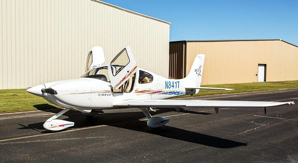 A white Cirrus SR20 aircraft with open doors and blue and red accent stripes parked on an airfield with hangars in the background.