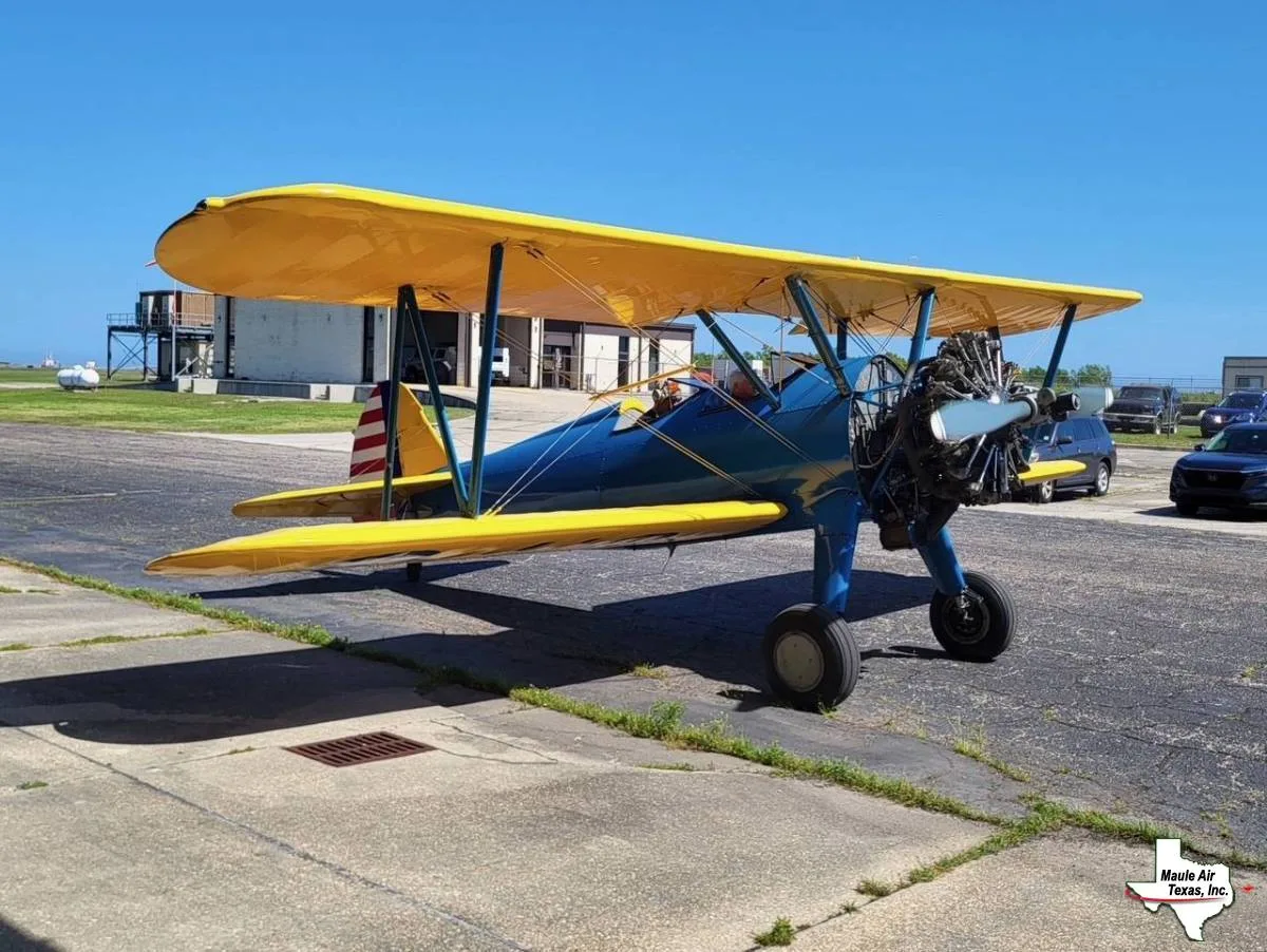 A blue and yellow biplane with an American flag tail is parked on an airfield tarmac under a clear blue sky.