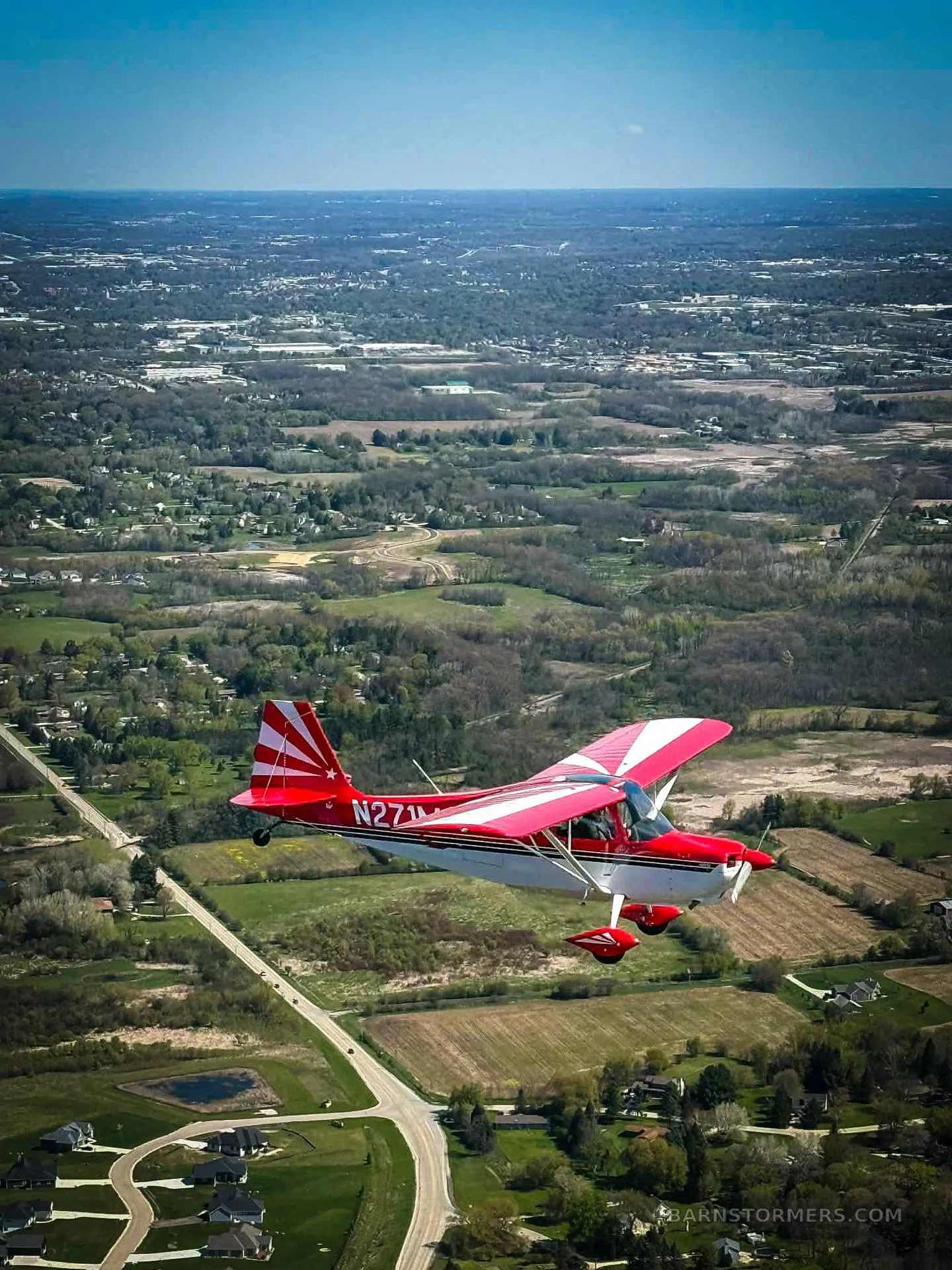 A red and white single-engine aircraft with tail number N271MA flies over a green rural landscape with scattered houses.