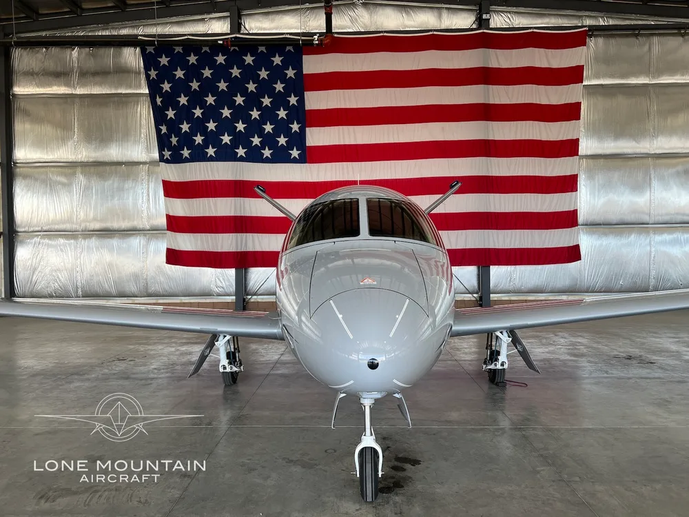 Front view of a grey and blue Cirrus Vision Jet (N33PK) parked in a hangar with an American flag in the background.