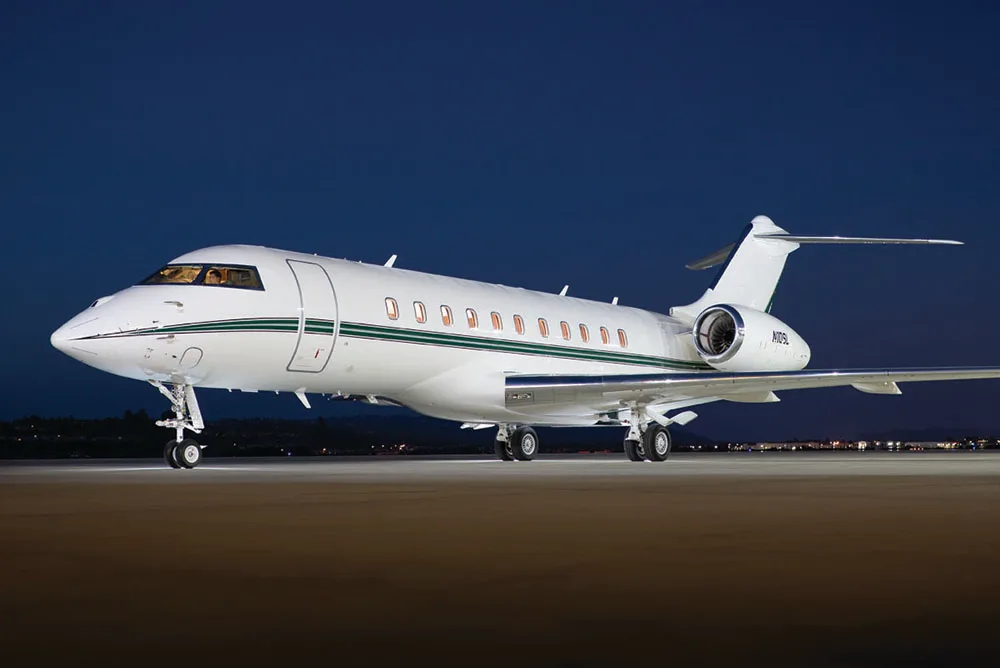 A white Bombardier Global Express business jet with a green stripe is parked on a tarmac at night, with city lights visible in the background.