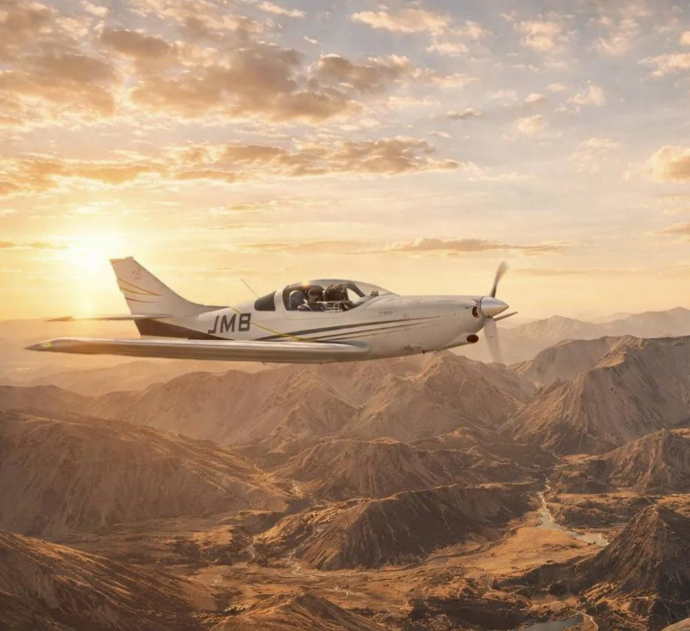A white JMB aircraft with gold and black accents flies over rugged mountains at sunset.