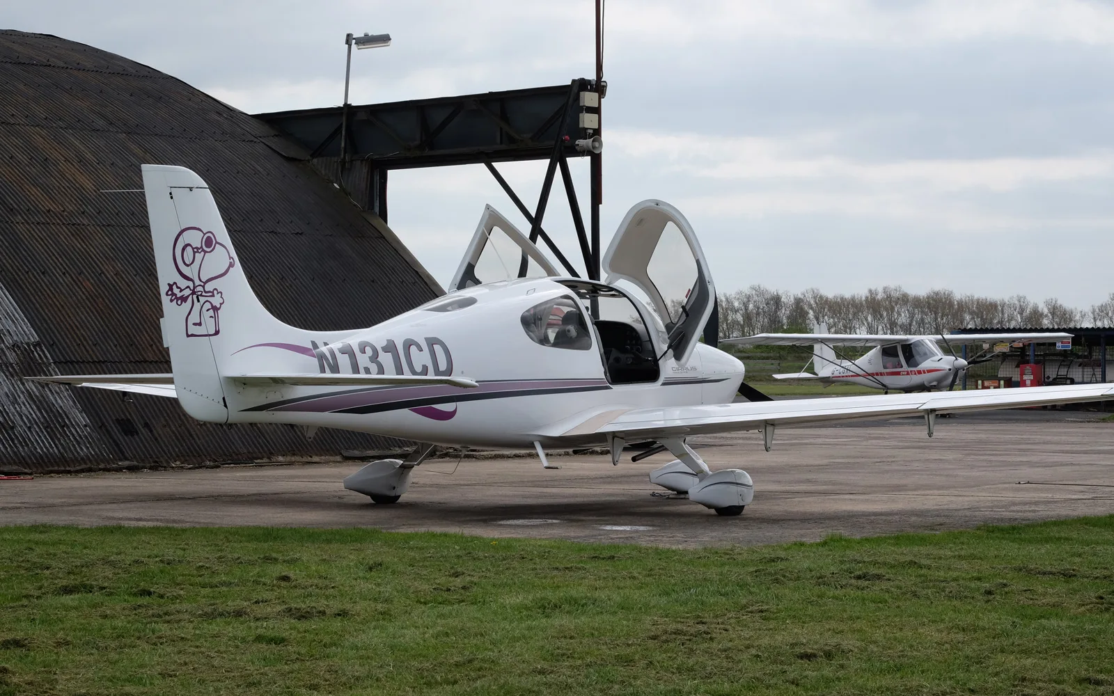 A white Cirrus SR22 aircraft with open gull-wing doors and a Snoopy graphic on the tail, parked on an airfield with another plane in the distance.