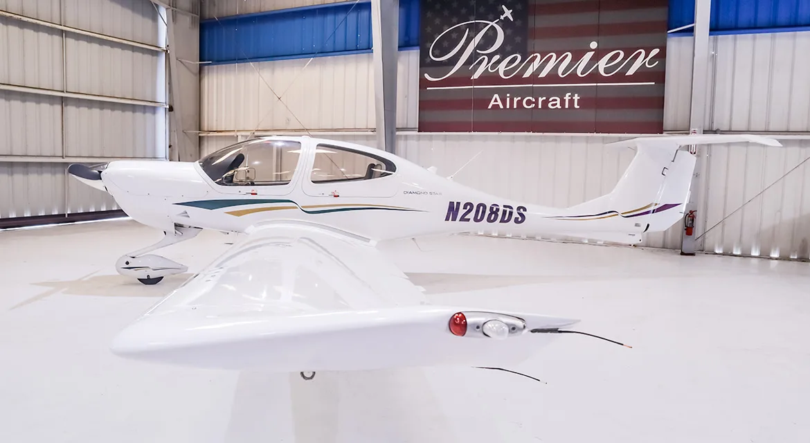 A white Diamond DA40 Star aircraft with green, gold, and purple stripes, registration N208DS, viewed from the front-right in a hangar.