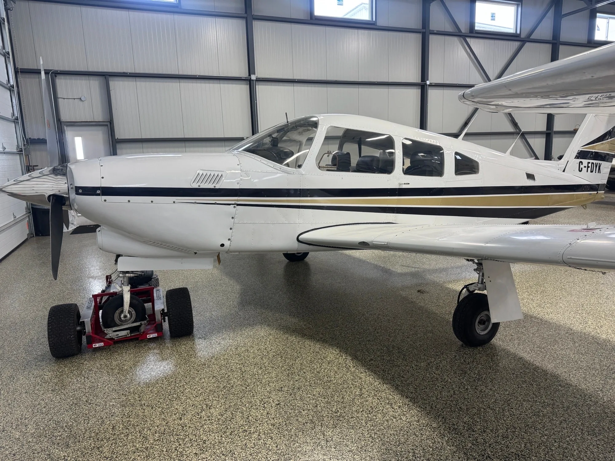 A white single-engine general aviation aircraft with black and gold stripes is parked in a modern hangar with an aircraft tug.