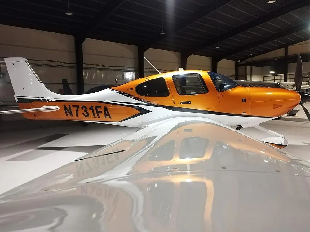 A side view of an orange and white Cirrus SR22 aircraft with registration N731FA inside a hangar, with a reflection on the wing.