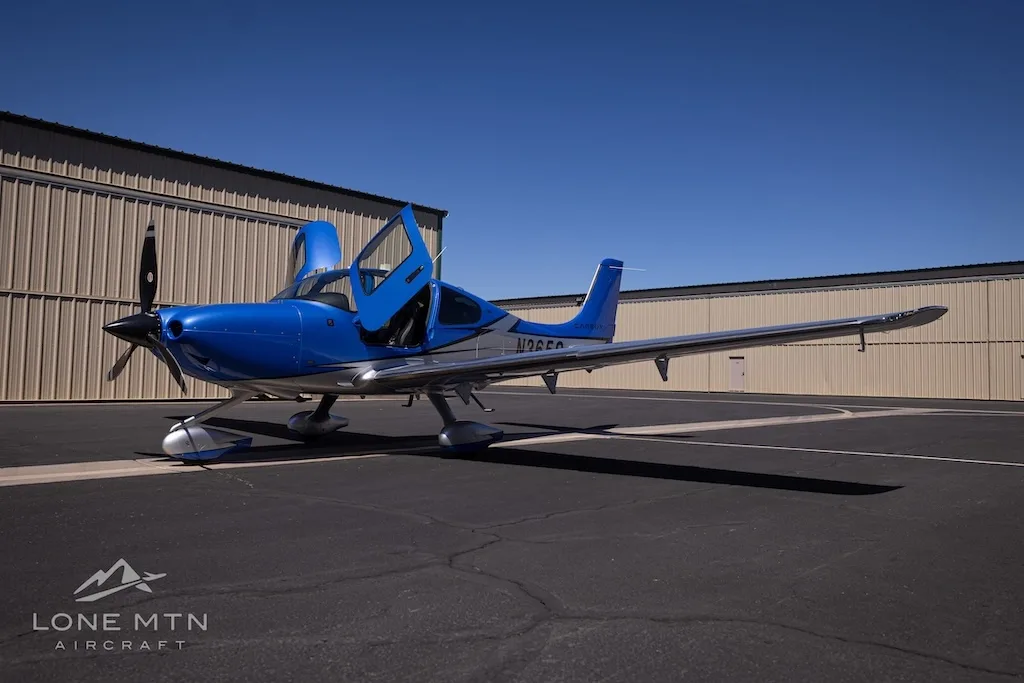 A blue and silver Cirrus SR22 aircraft, N365S, with its doors open, parked on an asphalt ramp in front of a hangar.