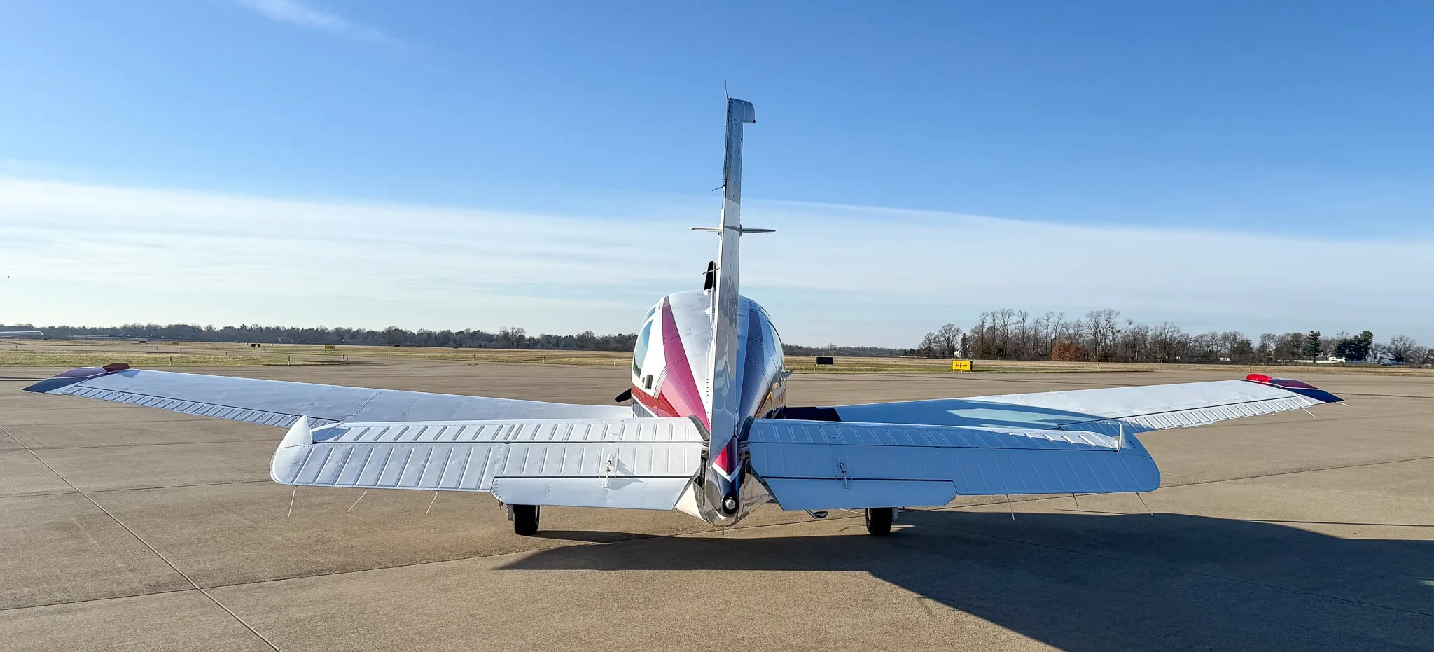 Rear view of a white Beechcraft Bonanza with red and blue tail stripes parked on an airfield.