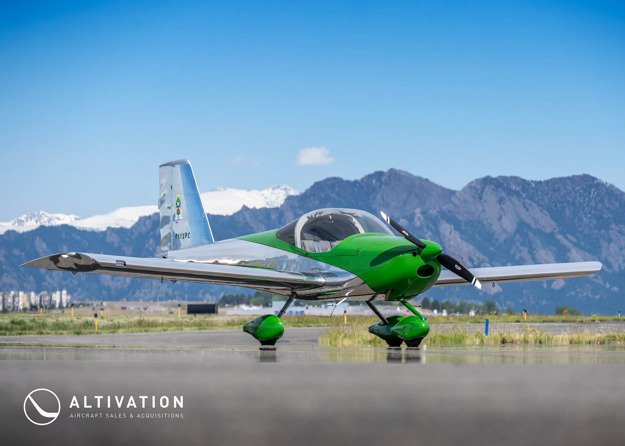 A low-angle front view of a green and polished aluminum Vans RV-12 aircraft, registration N612PC, on an airfield with snow-capped mountains.