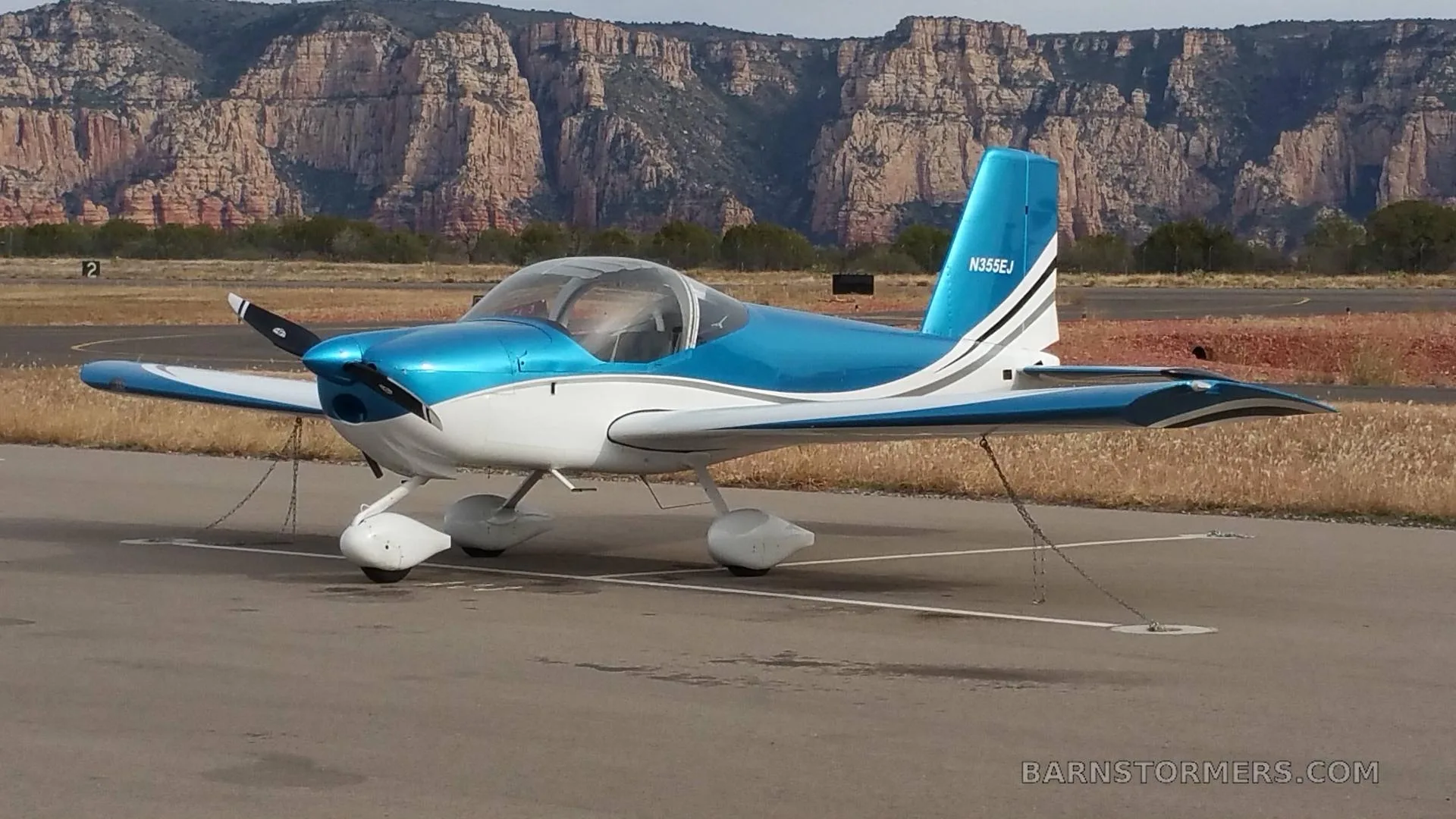 A blue and white single-engine aircraft with registration N355EJ is parked on an airfield with red rock mountains in the background.