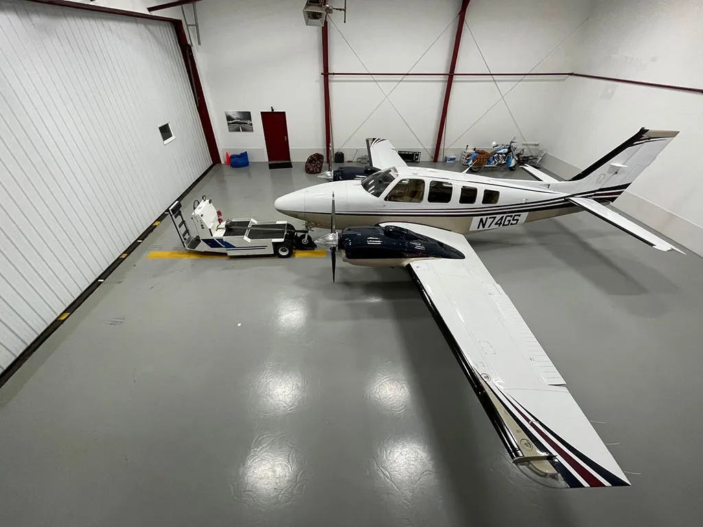 An overhead view of a white and tan twin-engine aircraft with dark blue accents parked in a hangar next to a tow vehicle.