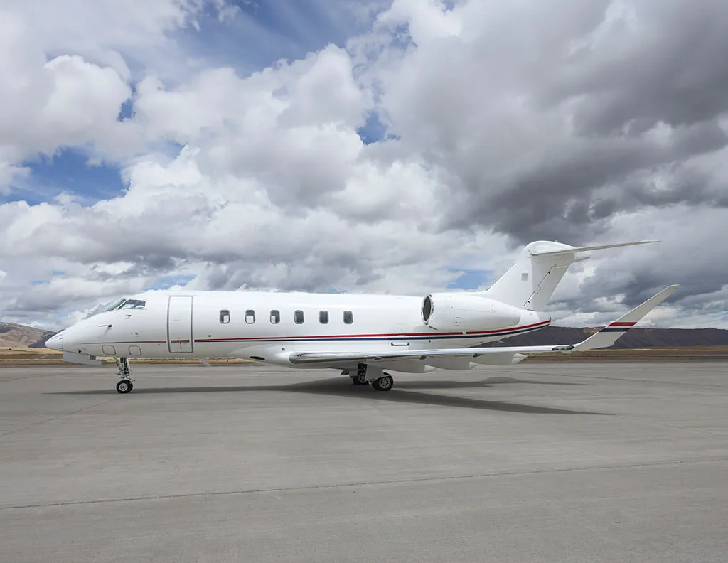 A white Bombardier Challenger 350 business jet with red and blue stripes on the tail, parked on an airfield under a partly cloudy sky.