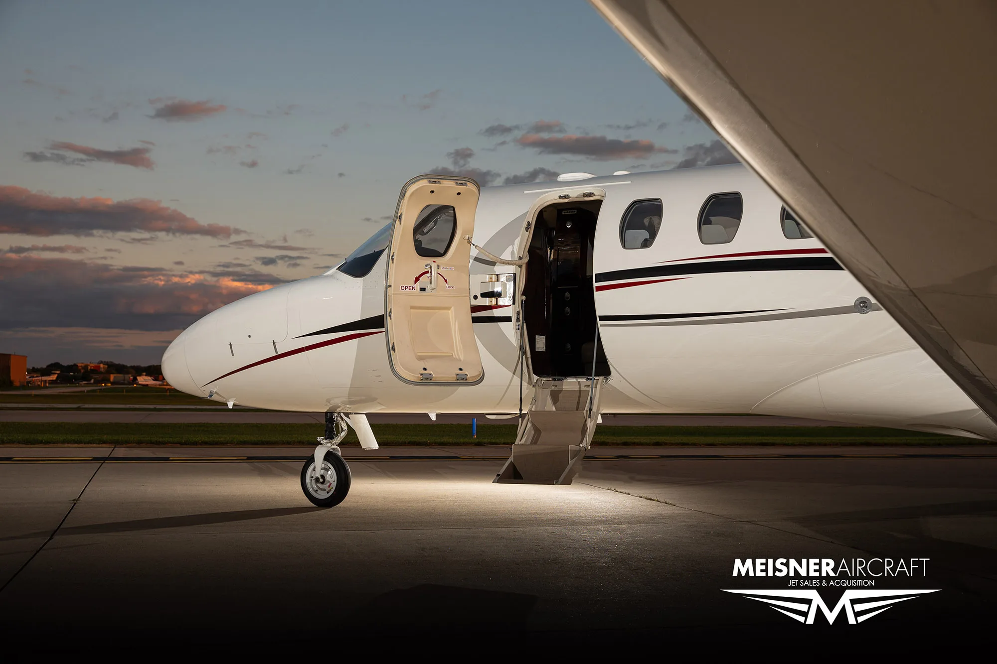 A close-up of the front fuselage of a white business jet with its main entry door open, illuminated on a tarmac at sunset.