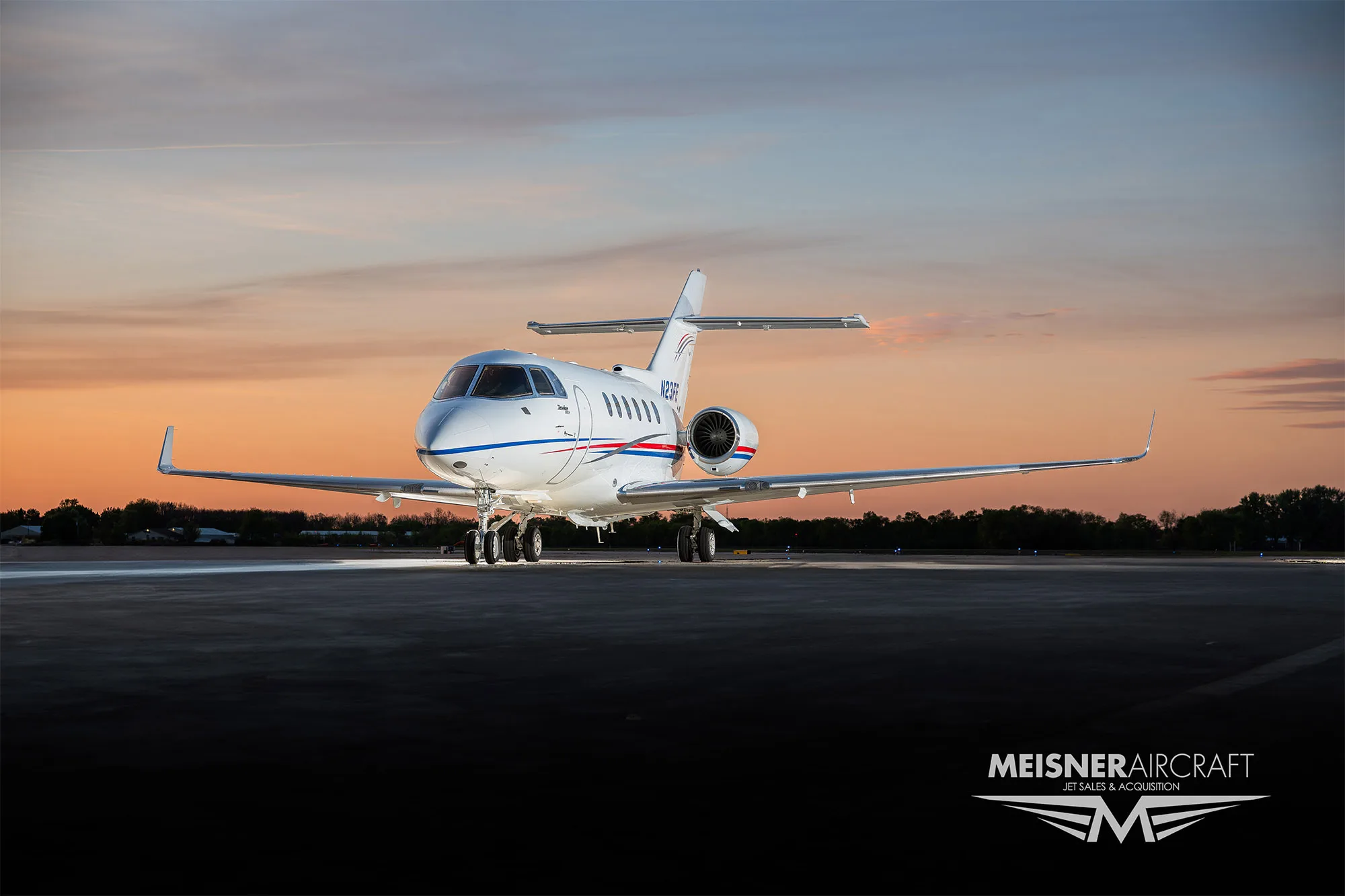 A white business jet with red and blue stripes, registration N23FE, parked on an airfield at sunset, viewed from the front.