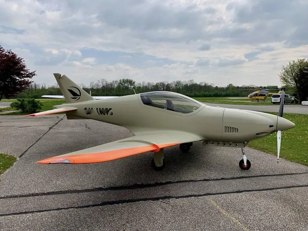 A light tan single-engine propeller aircraft with orange wingtips and tail markings, parked on an asphalt ramp under a cloudy sky.