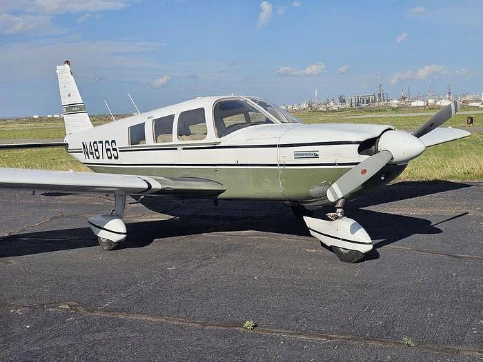 A white and green Piper Cherokee 180 aircraft (N4876S) is parked on an asphalt ramp with industrial buildings in the background.