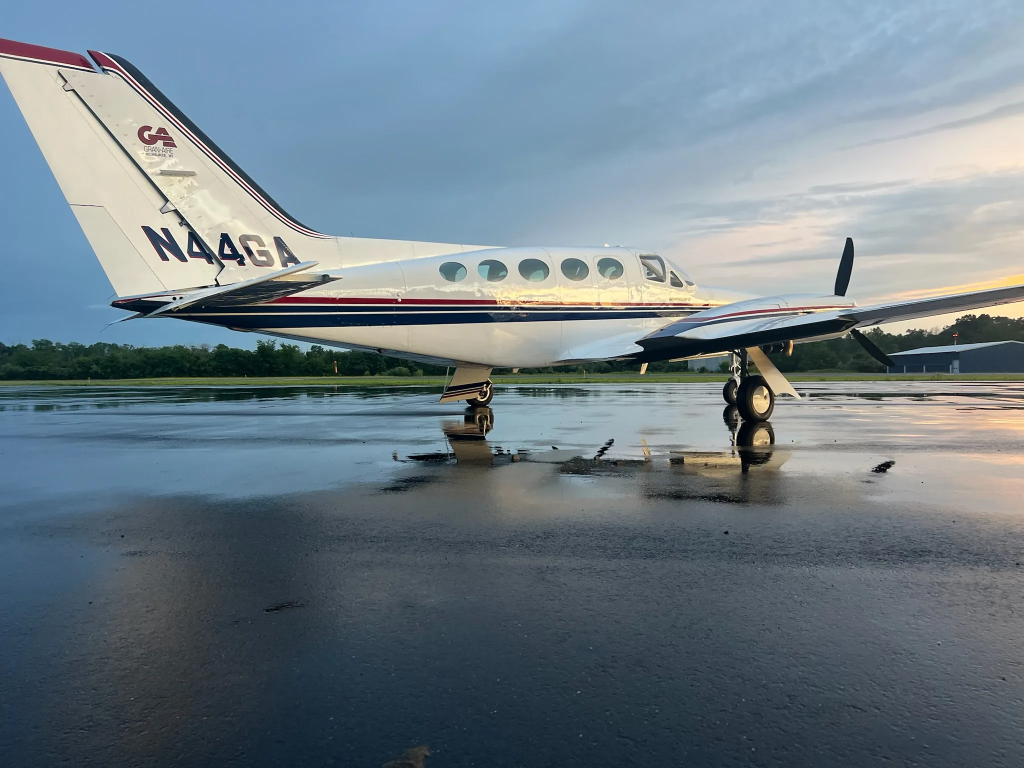 A white Cessna 414 Chancellor III with dark blue and red stripes parked on a wet tarmac at sunset.