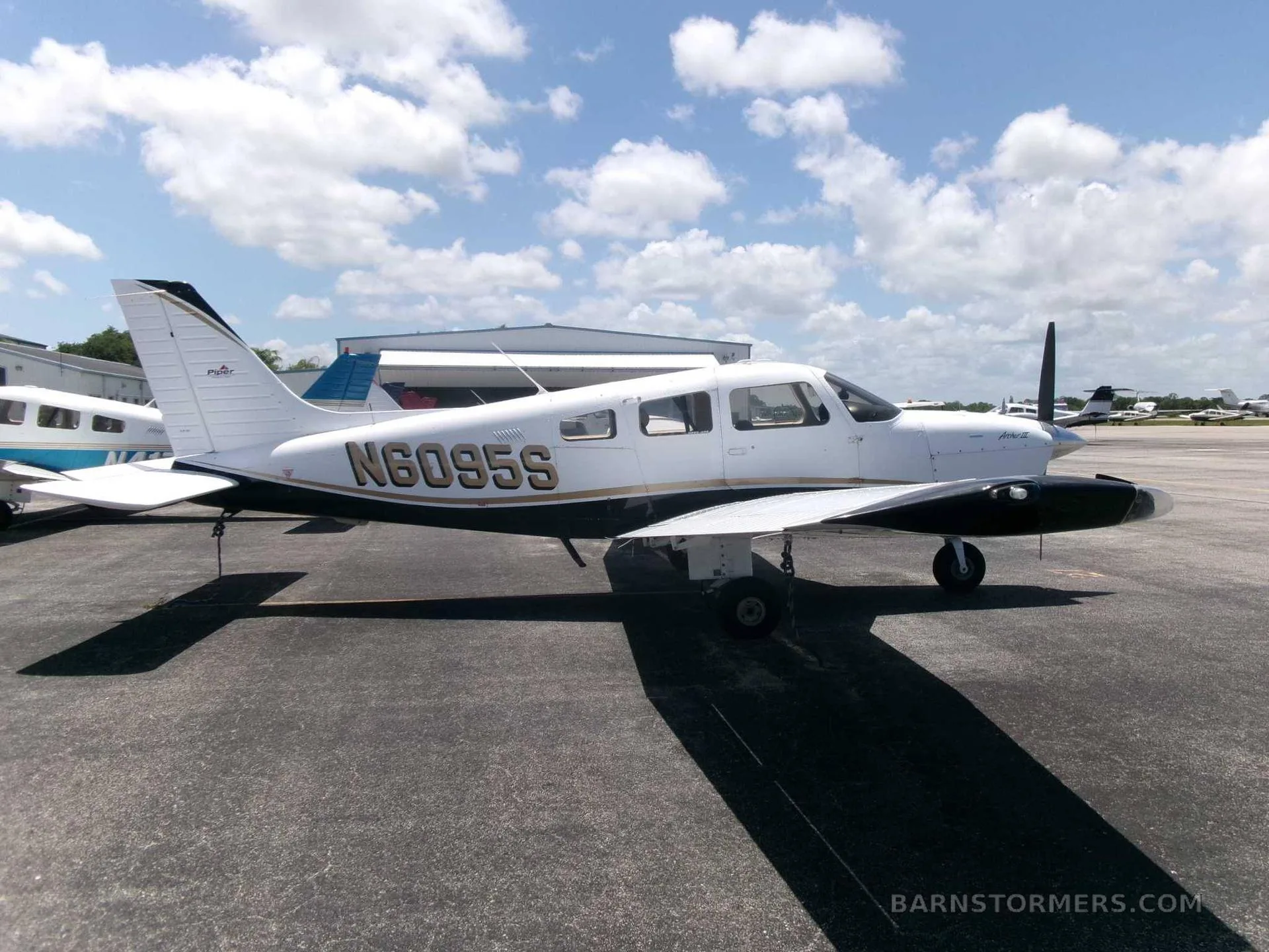 A white and black Piper PA-28 Cherokee with gold trim, registration N6095S, parked on an asphalt ramp with hangars in the background.