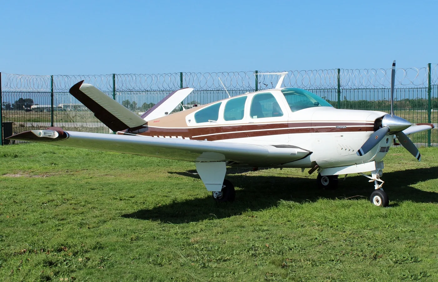 A white and brown single-engine propeller aircraft, likely a Beechcraft Bonanza, parked on a grassy field.