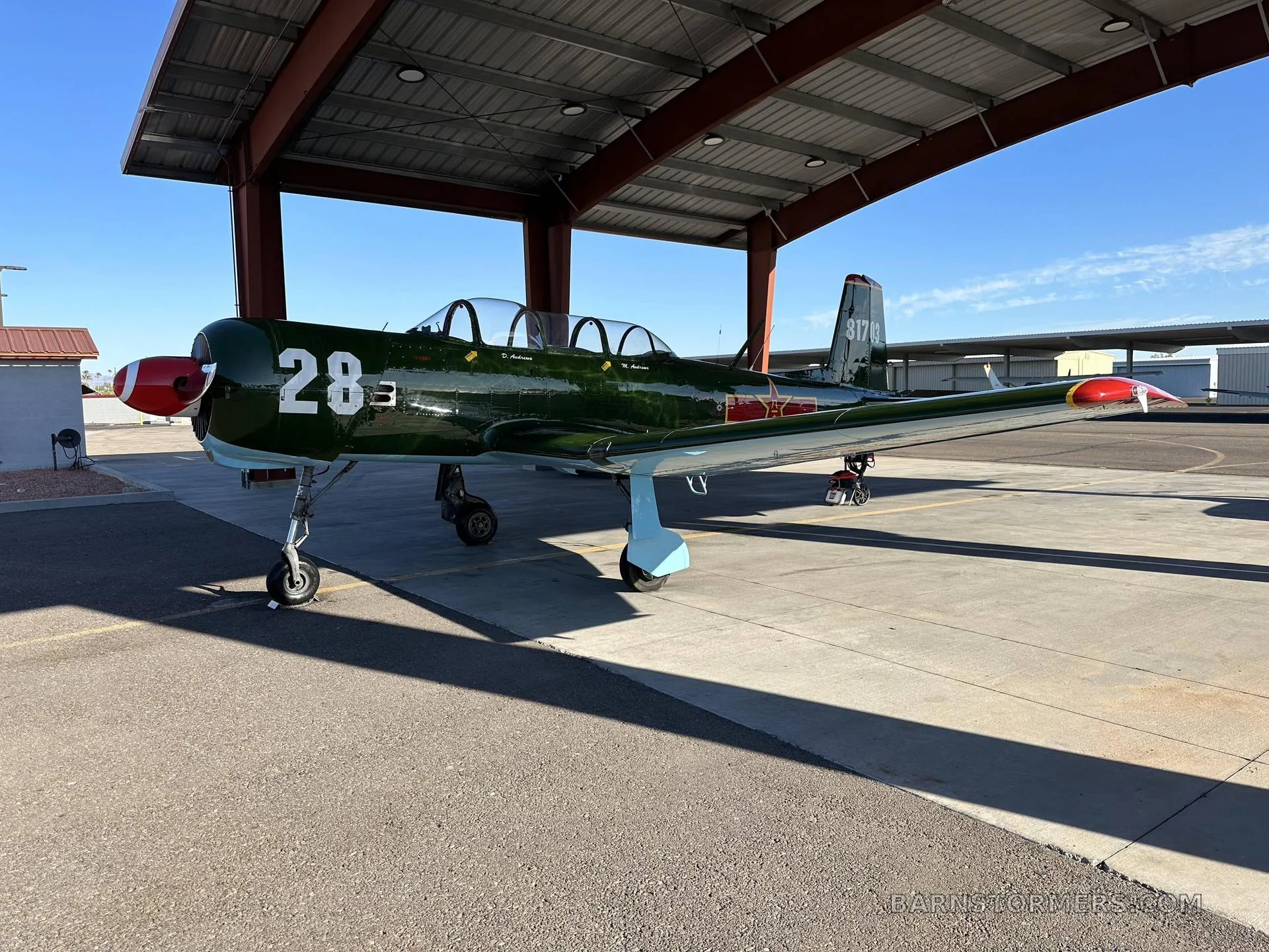 A green Nanchang CJ-6 aircraft with red nose and tail, number 28, parked under a hangar roof.