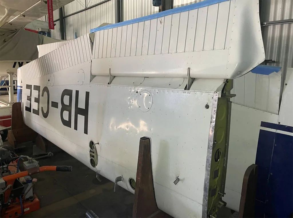 The white tail section of a Cessna F150 aircraft, registration HB-CEE, is seen disassembled in a hangar.
