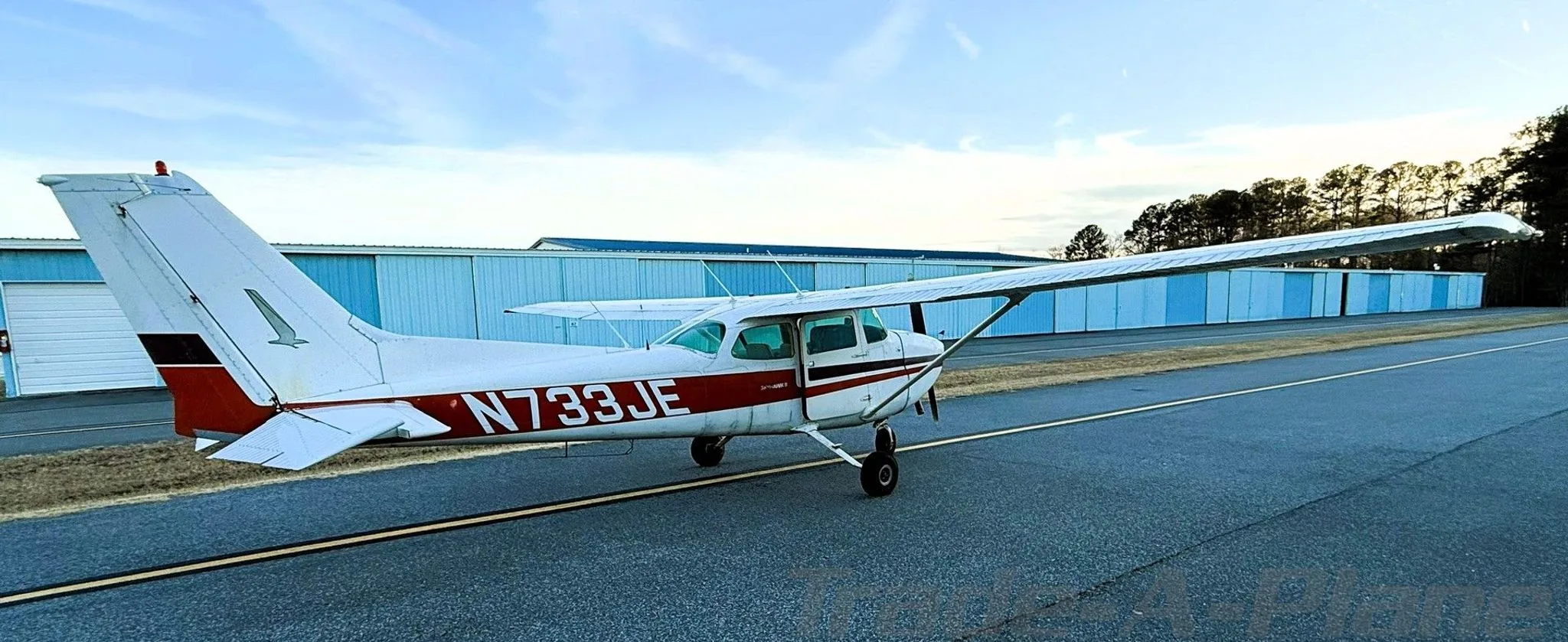 A white Cessna 172N with red and black stripes, registration N733JE, parked on an airfield with blue hangars in the background.