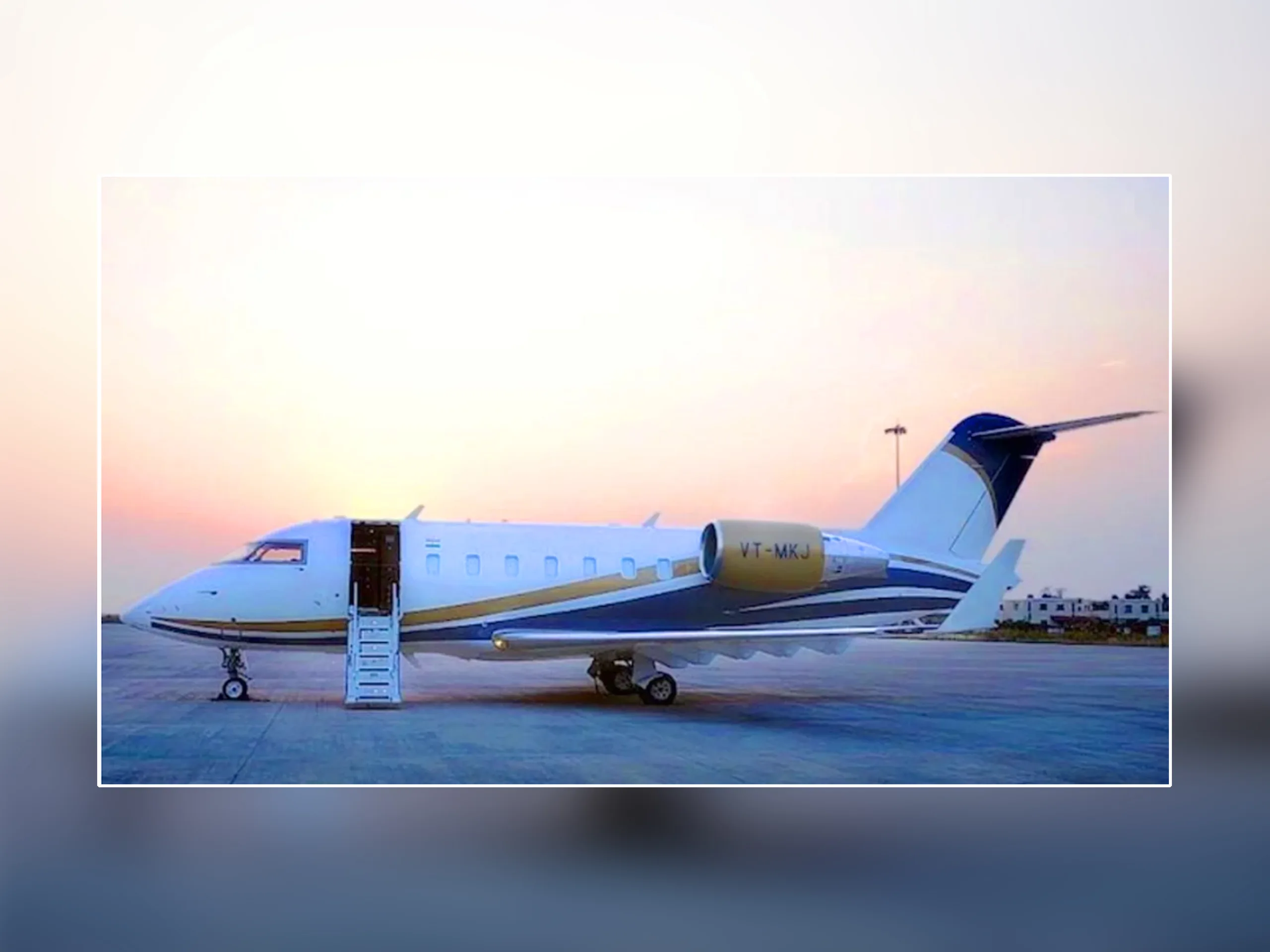 A white and blue Bombardier Challenger business jet with gold accents and registration VT-MKJ is parked on the tarmac at sunset with its airstairs deployed.