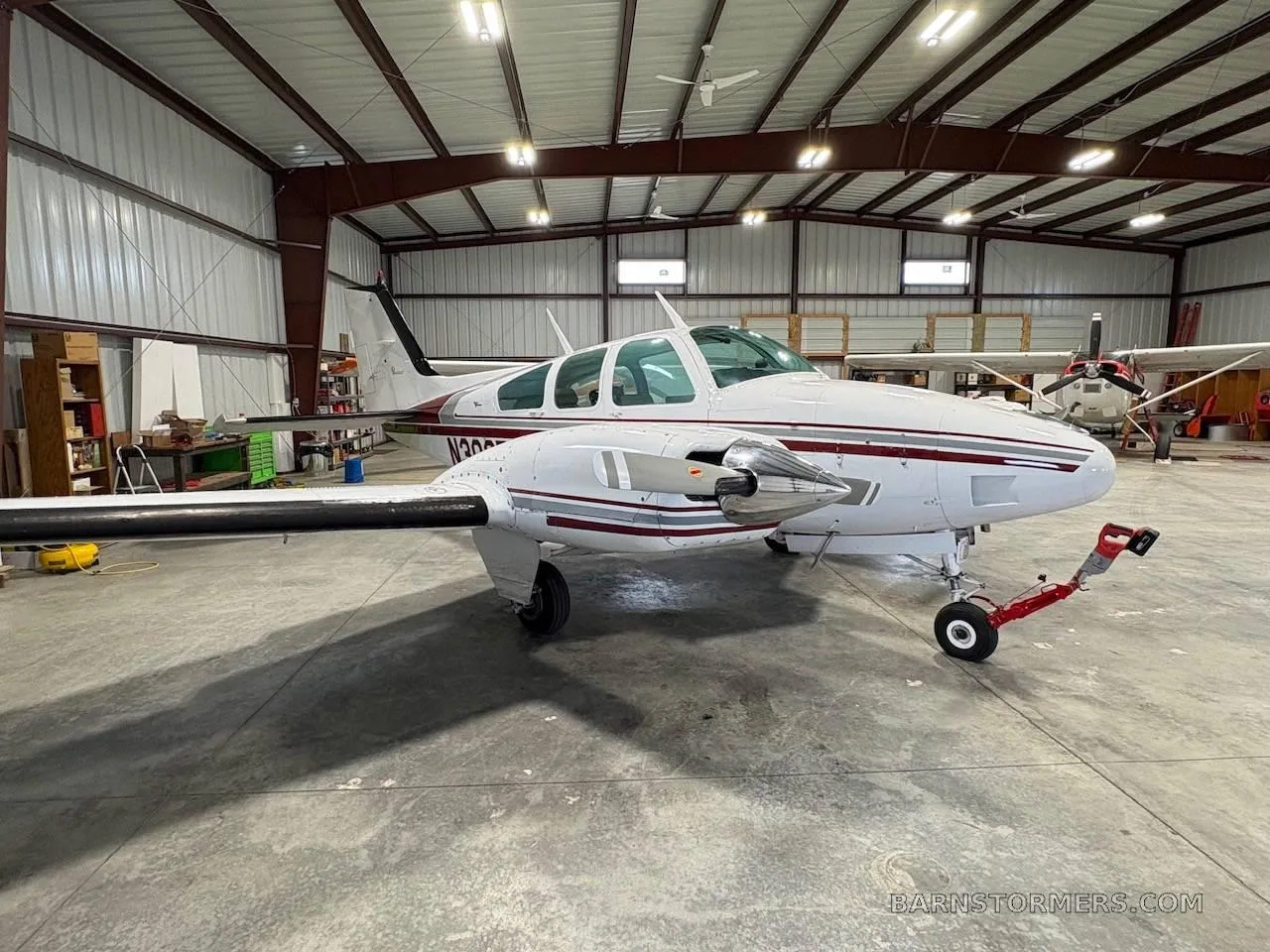 A white twin-engine propeller aircraft with red and grey stripes, seen from the front-right in a hangar.