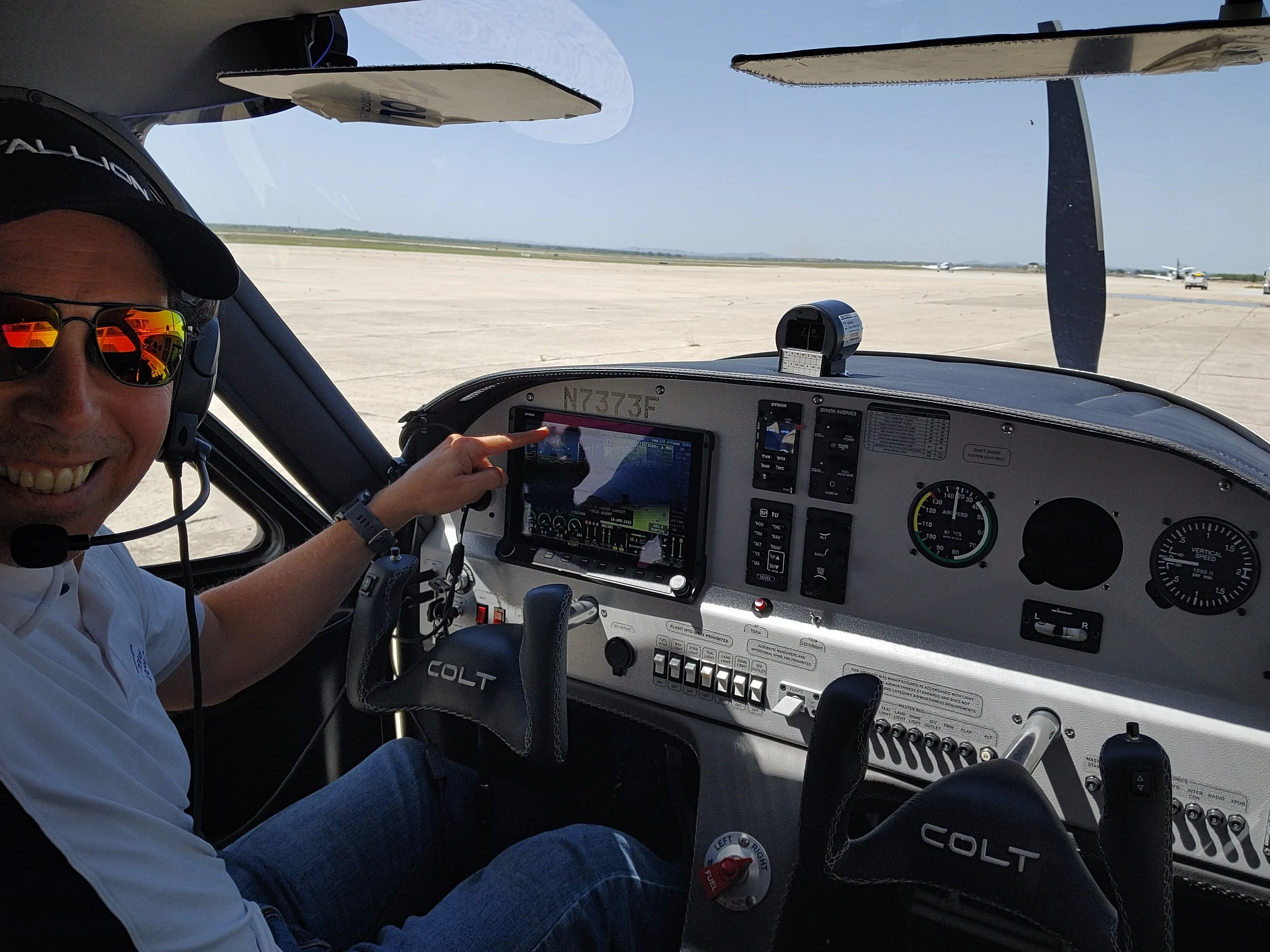 A pilot in the cockpit of a Texas Aircraft Colt N7373F pointing at the glass panel display on a sunny day at an airport.
