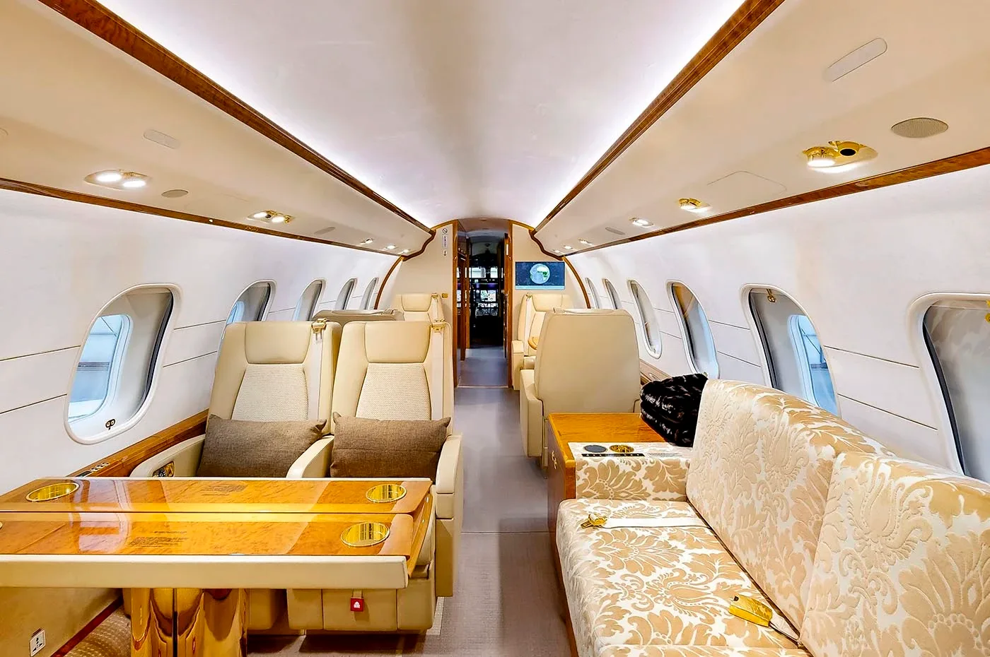The luxurious interior cabin of a private jet, featuring beige seating, a patterned sofa, and polished wood tables.